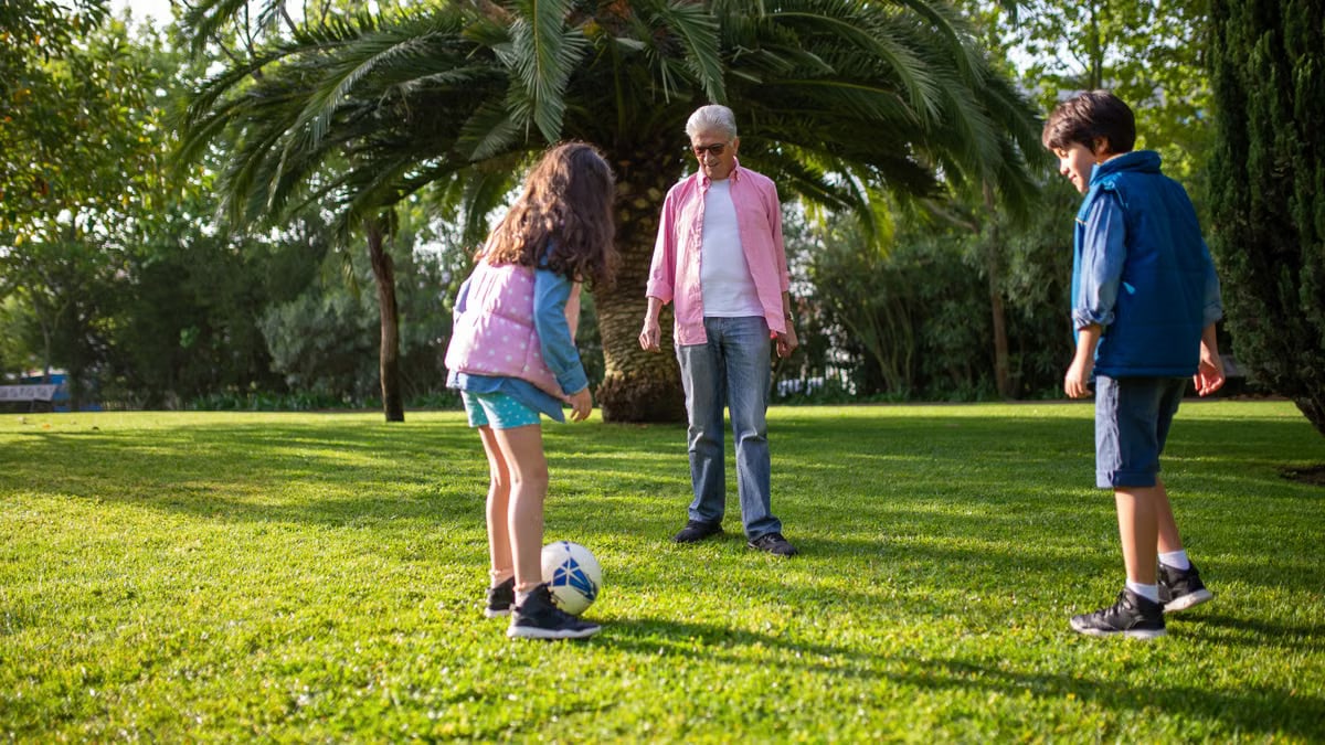 Grandfather and grandchildren playing soccer in a lush green park under a palm tree.